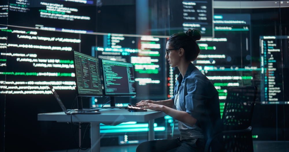 A female developer sitting at a desk with dual monitors filled with code, evaluating API integration needs for a project. Ideal for illustrating a decision-making checklist on whether to use an API in web or app development.