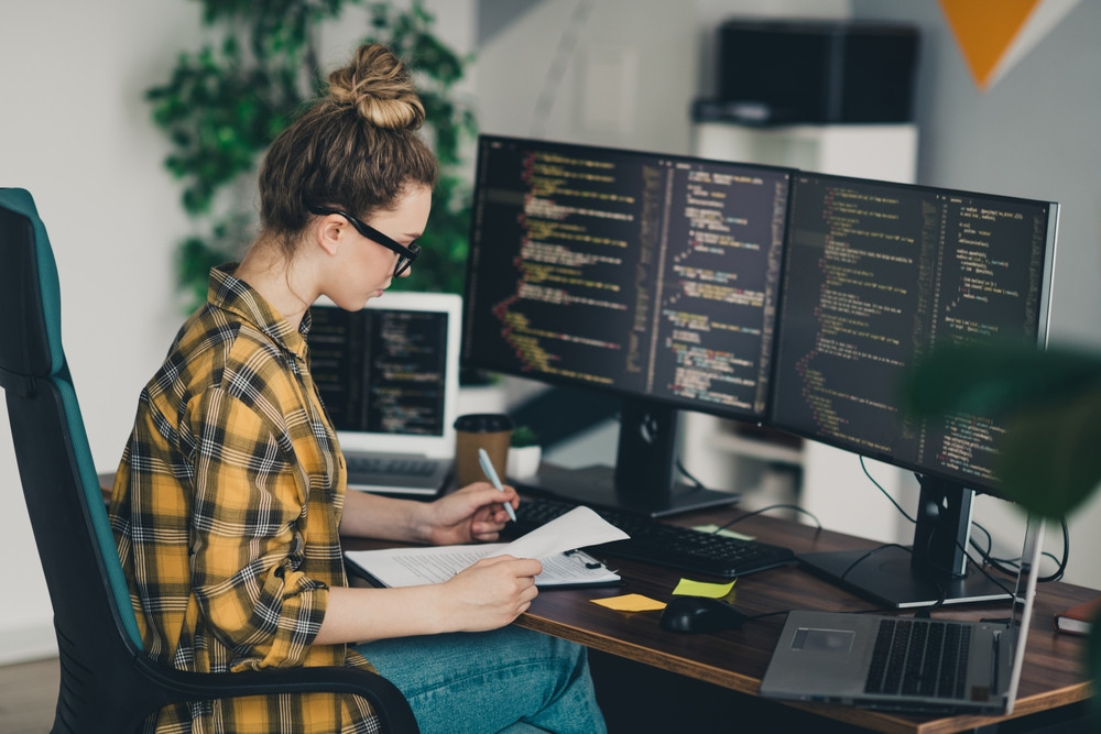 Young woman learning to code with multiple monitors displaying programming languages, representing beginner-friendly coding courses offered by Udemy and Coursera for HTML, Python, and JavaScript in 2026.