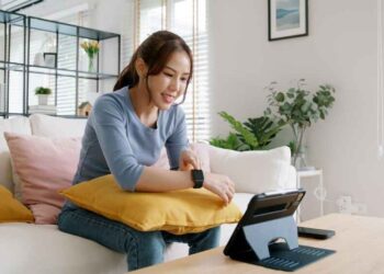 Young woman sitting on a couch at home checking her affordable fitness smartwatch connected to a tablet, demonstrating budget smartwatch tracking features like heart rate and workout monitoring.